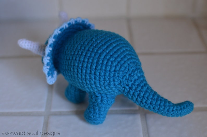 May include: A crocheted blue triceratops dinosaur toy with white accents. The toy has a frill around its head and a long tail.