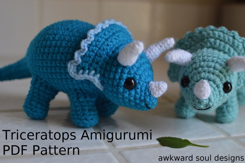 May include: Two crocheted amigurumi triceratops dinosaurs. One is teal blue and the other is a lighter blue. Both have white horns and eyes. The text "Triceratops Amigurumi PDF Pattern" is at the bottom of the image.