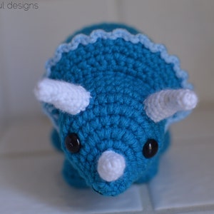 May include: A crocheted blue triceratops with white horns and a white frill. The triceratops has black eyes.