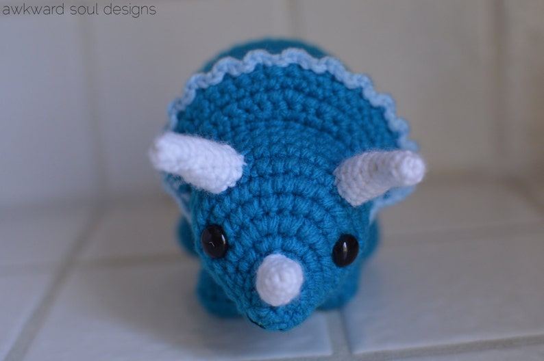 May include: A crocheted blue triceratops with white horns and a white frill. The triceratops has black eyes.