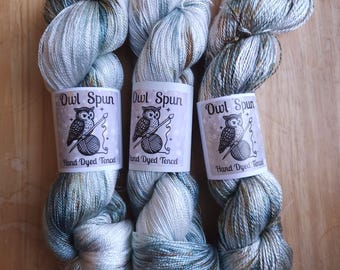 May include: Three skeins of hand-dyed Tencel yarn in shades of light blue, white, and brown. Each skein features a white paper label with the text Owl Spun and an illustration of an owl holding a knitting needle and a ball of yarn.