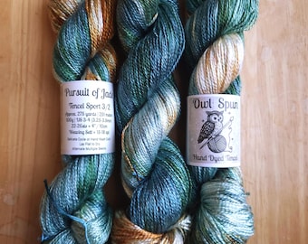 May include: Three skeins of Tencel Sport yarn in a variegated blend of teal, green, and golden brown. The left label reads Pursuit of Jade, Tencel Sport 3/2, 275 yards, 100g, US 3-4. The right label reads Owl Spun, Hand Dyed Tencel with an owl graphic.