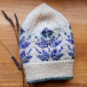 May include: A white knitted hat with a blue and purple floral pattern. The hat has a green trim around the bottom edge.