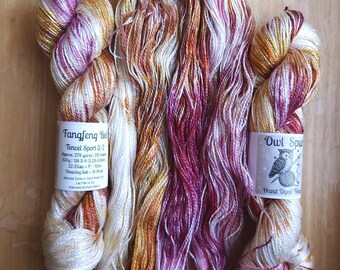 May include: Three skeins of hand-dyed Tencel sport yarn featuring a variegated color palette of deep magenta, golden orange, and cream white. Two skeins have paper labels, one reading Fangfeng Bell and the other Owl Spun Hand Dyed.