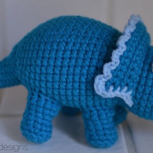 May include: A crocheted blue triceratops dinosaur toy with white horns and a light blue frill.