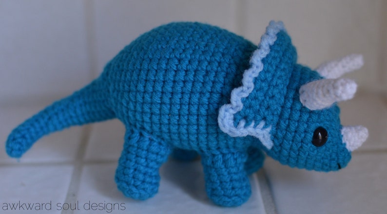 May include: A crocheted blue triceratops dinosaur toy with white horns and a light blue frill.