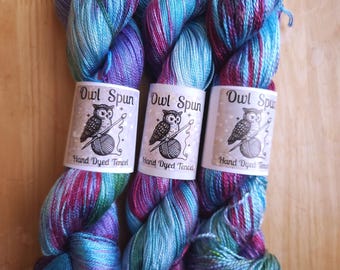 May include: Three skeins of hand-dyed Tencel yarn featuring a variegated color palette of bright blue, purple, and magenta. Each skein is wrapped in a white paper label with the text Owl Spun and an illustration of an owl with yarn.