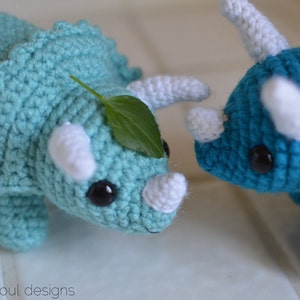 May include: Two crocheted dinosaur toys. One is teal blue with a green leaf on its head and the other is a darker blue.