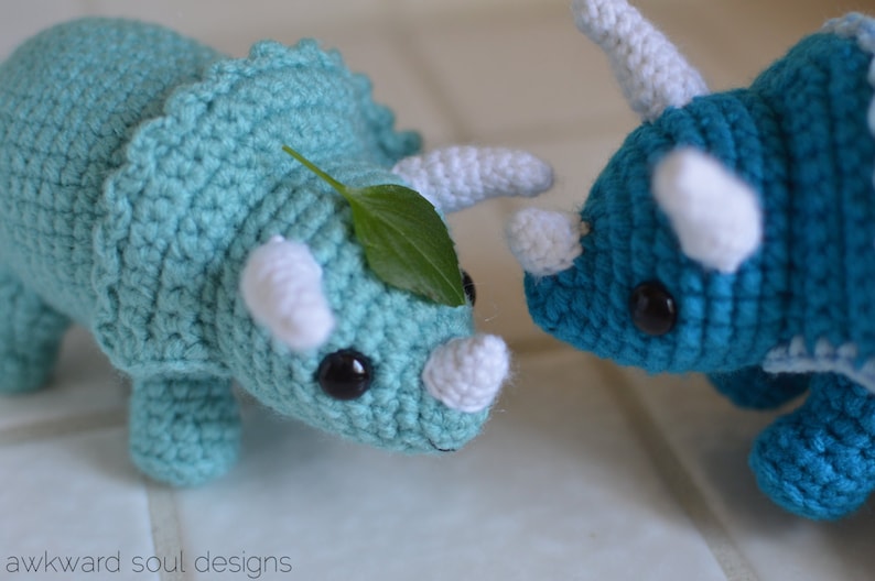May include: Two crocheted dinosaur toys. One is teal blue with a green leaf on its head and the other is a darker blue.