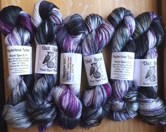May include: Six skeins of hand-dyed Tencel yarn arranged in a row, featuring a variegated color palette of black, white, gray, and deep purple. Each skein has a paper label with text including Mysterious Lotus or Owl Spun branding.