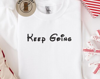 May include: White crew neck sweatshirt with the phrase "Keep Going" embroidered in black script. The sweatshirt has a classic fit and is made of a soft material. A circular "Embroidery" logo is visible in the upper left corner.