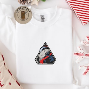 以下が含まれることがあります： 黒い持ち手と「But First, Coffee」のテキストが黒い太字で書かれた白いセラミックマグ。マグカップには濃い液体、おそらくコーヒーが入っており、明るい色の表面に置かれています。