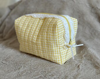 May include: A yellow and white gingham makeup bag with a white lace trim and a zipper closure. The bag has a white ribbon tied around the zipper pull.