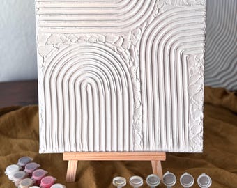 May include: A white canvas painting with raised lines and arches, on an easel, with a set of paint pots and brushes in front of it.