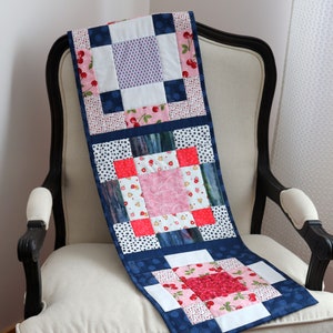 May include: A blue and white patchwork table runner with a red and pink floral pattern. The runner is made of squares and has a quilted texture.