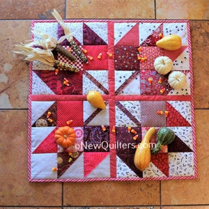 May include: A square quilted table runner with a patchwork design in shades of red, white, and burgundy. The runner is decorated with small pumpkins, gourds, and candy corn. The edge is trimmed with a pink and white rope-like border. The text "oNewQuilters.com" is visible.