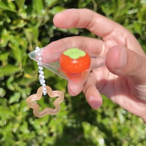 May include: A clear plastic keychain with a small orange and green persimmon charm attached to a silver chain and a clear star-shaped charm.