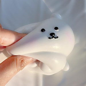 May include: A white, seal-shaped toy with black eyes and a black nose. The toy has a smooth surface and is being held by a hand. The seal has a pink blush on its cheeks.