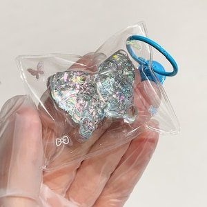 May include: A clear plastic keychain with a silver iridescent butterfly charm. The keychain has a blue ring and a small bow and butterfly design on the plastic.