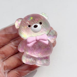 May include: A holographic pink teddy bear figurine with a light pink bow tie. The bear has black eyes and a white snout. The figurine is held in a gloved hand against a white background.