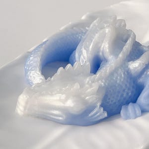 以下が含まれることがあります： 青と白のドラゴンの彫刻で、尻尾が丸まっており、寝ているポーズをとっています。ドラゴンは滑らかで光沢のある素材で作られています。