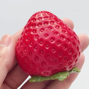Pode incluir: Um objeto em forma de morango vermelho brilhante com uma superf&iacute;cie texturizada apresentando pequenas reentr&acirc;ncias no exterior. O objeto tem uma pequena estrutura verde em forma de folha na base, imitando a apar&ecirc;ncia de um morango real.