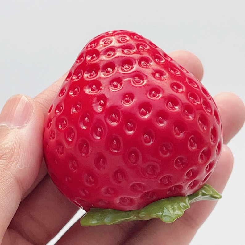 Pode incluir: Um objeto em forma de morango vermelho brilhante com uma superf&iacute;cie texturizada apresentando pequenas reentr&acirc;ncias no exterior. O objeto tem uma pequena estrutura verde em forma de folha na base, imitando a apar&ecirc;ncia de um morango real.