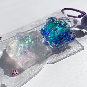 May include: Two resin flower charms in a clear plastic bag. One charm is iridescent white and the other is blue with green glitter.