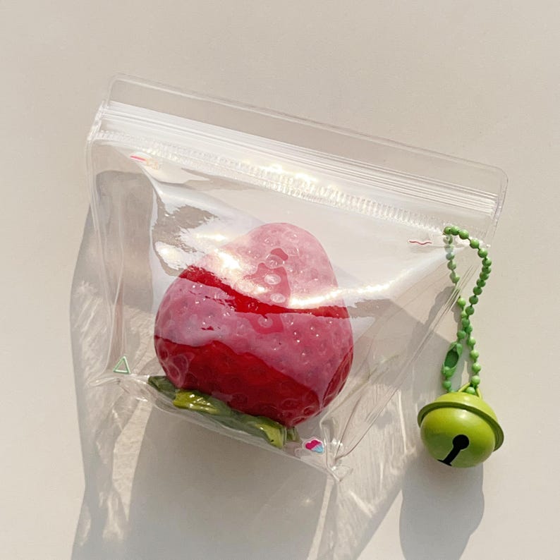 Pode incluir: Uma bolsa de pl&aacute;stico transparente com fecho de correr contendo um objeto vermelho em forma de morango com uma base de folhas verdes. Uma corrente de esferas verde com um pequeno sino verde est&aacute; presa &agrave; lateral da bolsa.