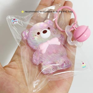 May include: A clear star-shaped PVC bag containing a glittery iridescent purple bear figure with a white muzzle and a pink bow. The bag features the text recommend to squeeze it and Lucky Bear, with a pink ring and a small pink bell attached to the top.