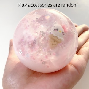 May include: A pink, spherical decorative item with a cartoon cat on an ice cream cone inside. The sphere is filled with glitter and star-shaped confetti. Text at the top reads "Kitty accessories are random."