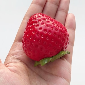 Pode incluir: Uma r&eacute;plica de morango vermelho brilhante com um acabamento lustroso e uma superf&iacute;cie texturizada com pequenas reentr&acirc;ncias. O fruto tem uma pequena estrutura de folha verde moldada na base, conferindo um aspeto realista a este objeto decorativo.