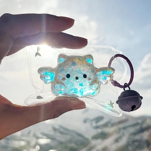 以下が含まれることがあります： 小さな羽のついたクマの形をした、紫色のベルと紫色のリングが付いた、透明で虹色のキーホルダーチャーム。クマは青く光り、明るい空を背景に持っています。