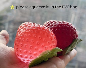 Könnte beinhalten: Zwei erdbeerförmige Quetschspielzeuge mit grünen Kunststoffblättern. Eine Erdbeere ist durchscheinend hellrot, die andere undurchsichtig dunkelrot. Der Text "⭐ please squeeze it in the PVC bag" ist über den Objekten zu sehen.