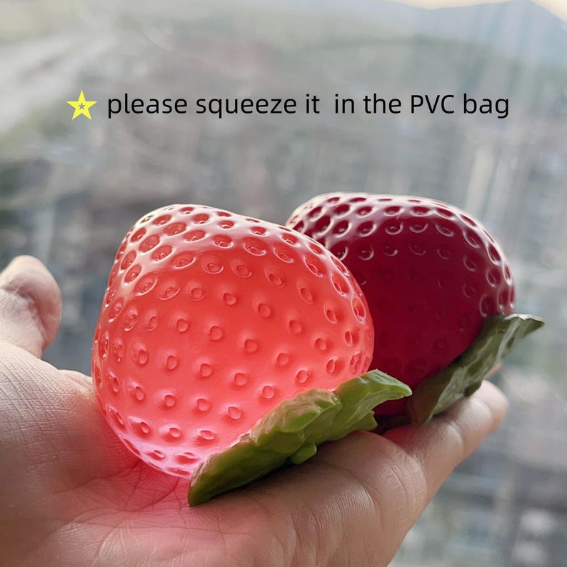Pode incluir: Dois brinquedos de apertar em forma de morango com folhas de pl&aacute;stico verde. Um morango &eacute; vermelho claro transl&uacute;cido e o outro &eacute; vermelho escuro opaco. O texto "⭐ please squeeze it in the PVC bag" &eacute; vis&iacute;vel acima dos objetos.