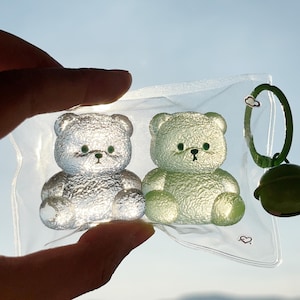 Op de afbeelding: Twee doorschijnende teddybeerbeeldjes, één zilver en één groen, worden in een doorzichtige plastic zak gehouden. De beren hebben zwarte ogen en neuzen. Een groene sleutelhanger met een wolkbedel en een groene bel is zichtbaar.