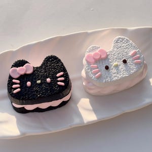 May include: Two decorative Hello Kitty-shaped items, one black and one white, sit on a white platter. The black item has a pink bow and accents, while the white one has a pink bow and pink accents. Both have a pink base.