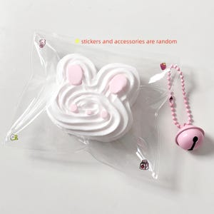 May include: A white, swirl-textured, rabbit-shaped object inside a clear plastic bag with small printed icons. The text "stickers and accessories are random" is printed on the bag. A pink ball-chain keychain with a bell is attached to the side.
