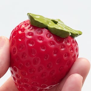 Pode incluir: Uma figura de morango vermelho brilhante com uma superf&iacute;cie texturizada apresentando pequenas reentr&acirc;ncias e um acabamento brilhante. O topo do morango &eacute; adornado com um elemento de folha verde s&oacute;lido e esculpido sobre a fruta.