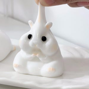 May include: A white, squishy, hamster-shaped toy with black eyes. The toy is being squeezed by a hand.