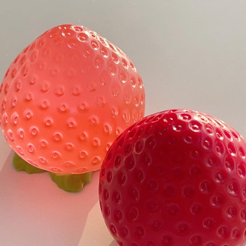 Pode incluir: Dois objetos esf&eacute;ricos em forma de morango com superf&iacute;cies texturizadas e com covinhas. Um objeto &eacute; de cor rosa coral claro transl&uacute;cido, enquanto o outro &eacute; de um vermelho profundo s&oacute;lido e opaco. Ambos os itens possuem uma base de folha verde moldada.