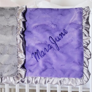 May include: A soft, textured blanket featuring a dual-color design with one side in grey and the other in purple. The purple section is embroidered with the name Mara June in cursive script. Both sides are finished with a ruffled satin border.