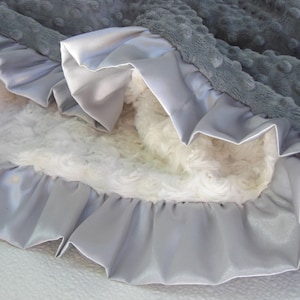 May include: A gray and white baby blanket with a satin ruffle trim. The blanket is soft and cuddly, perfect for a baby's nursery.