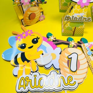May include: A yellow and black bee-themed birthday decoration set. Includes a bee, a rainbow, a flower, a honeycomb with the number 1, and the name "Ariadne". The set is on a yellow surface.