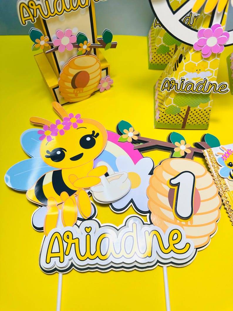 May include: A yellow and black bee-themed birthday decoration set. Includes a bee, a rainbow, a flower, a honeycomb with the number 1, and the name "Ariadne". The set is on a yellow surface.