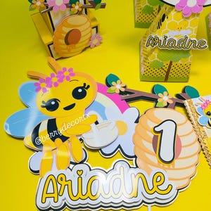 May include: A yellow bee-themed birthday party decoration set. Includes a bee-shaped figure with flower accents, a honeycomb, and the name "Ariadne" on a cake topper. The number "1" is also featured.