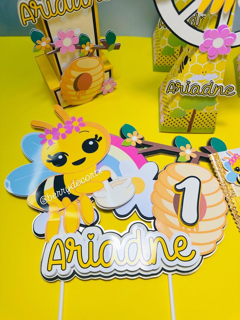 May include: A yellow bee-themed birthday party decoration set. Includes a bee-shaped figure with flower accents, a honeycomb, and the name "Ariadne" on a cake topper. The number "1" is also featured.