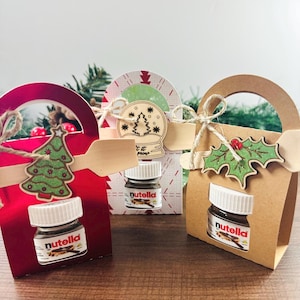 Peut inclure: Trois petits sacs cadeaux avec des pots de Nutella miniatures. Les sacs sont rouges, blancs et marron, décorés de découpes sur le thème de Noël : un sapin, une boule à neige et du houx. Chaque pot a un couvercle blanc et une étiquette Nutella.