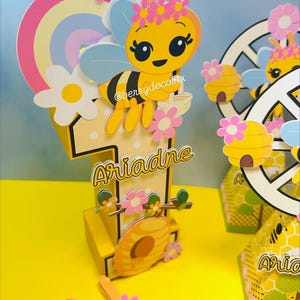 May include: A yellow and pink bee-themed birthday decoration featuring a large number one with the name "Ariadne" in gold script. The design includes a cartoon bee, flowers, a rainbow, and a beehive. The image also shows additional matching party favors.