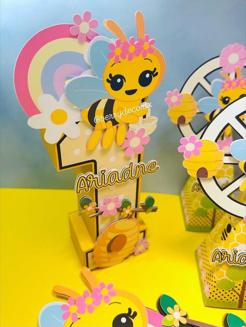 May include: A yellow and pink bee-themed birthday decoration featuring a large number one with the name "Ariadne" in gold script. The design includes a cartoon bee, flowers, a rainbow, and a beehive. The image also shows additional matching party favors.